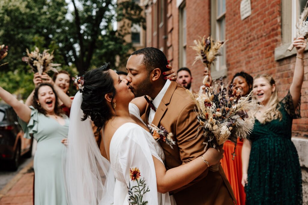 An interracial newlywed couple embrace in a kiss in front of their wedding party, who is cheering behind them. There are brick buildings and trees in the background.