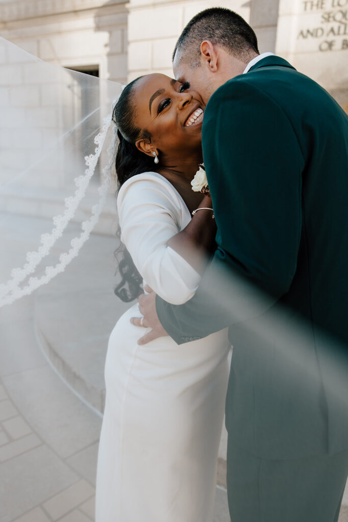 A groom kisses a bride on her cheek while the bride smiles. Her veil is blowing in the wind in front of the camera.