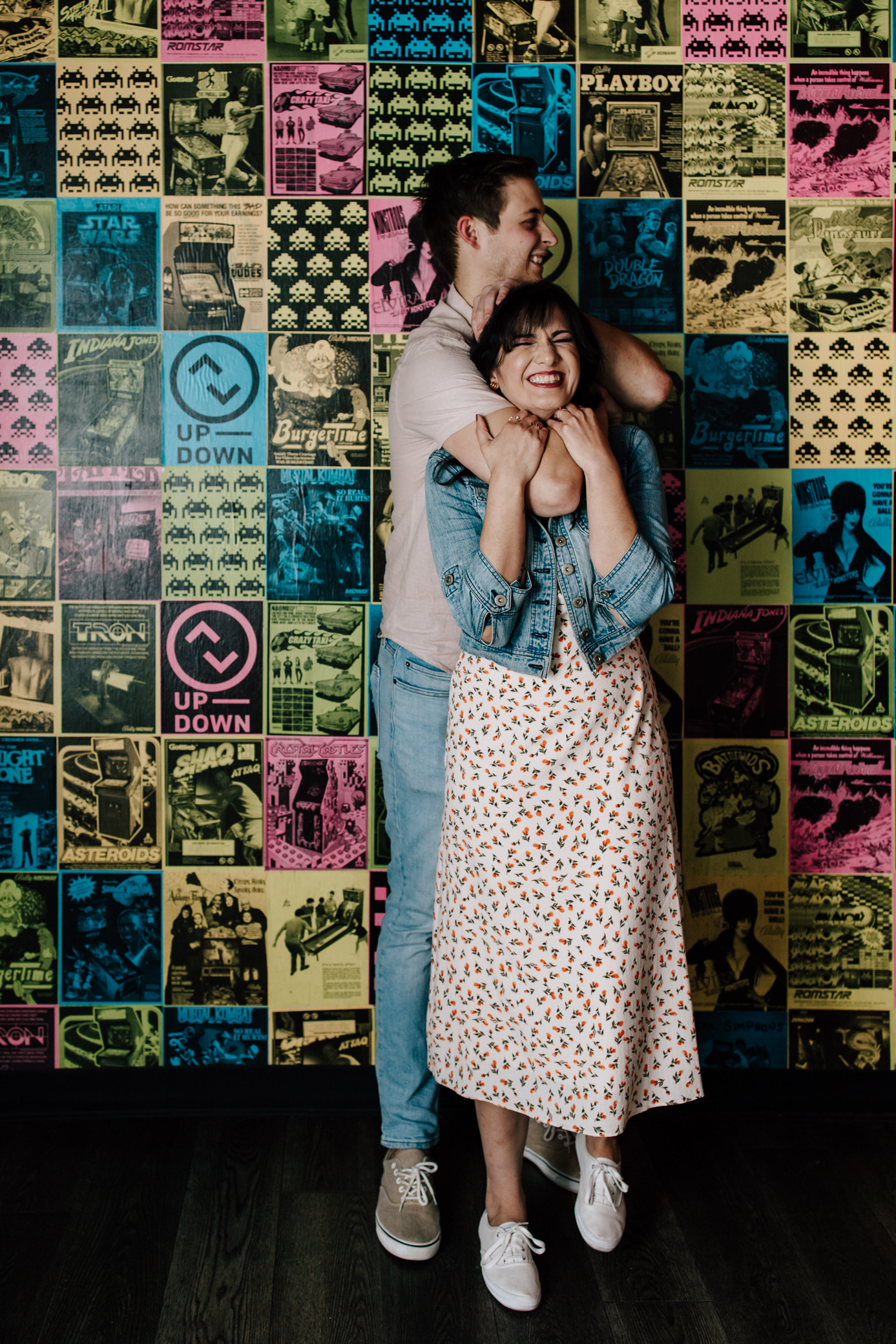 An engaged couple standing in front of a wall with arcade graphics at Up-Down Arcade in St. Louis, Missouri. The man playfully has the woman in a chokehold and they're both smiling.