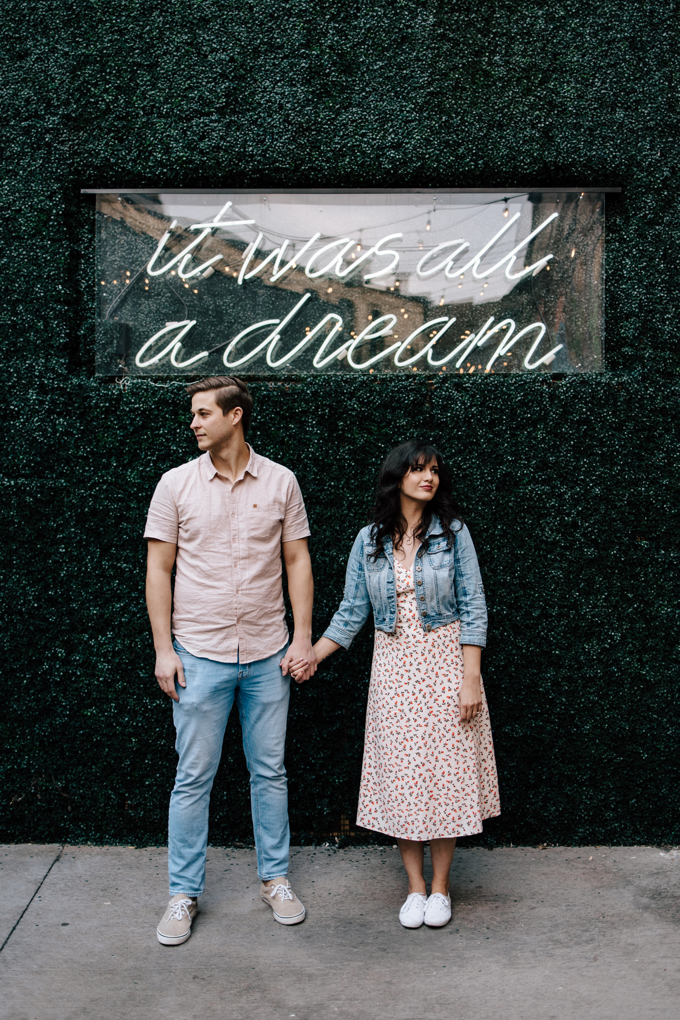 A man and woman hold hands looking in opposite directions under a sign attached to a greenery wall that reads "it was all a dream."
