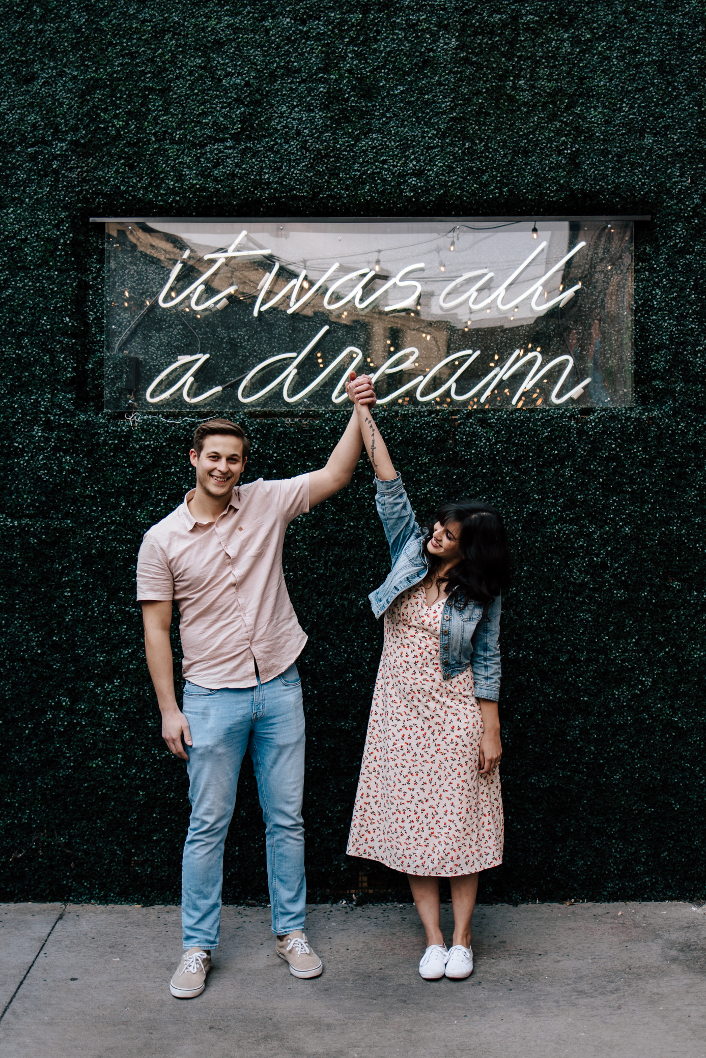 A man and woman stand under a sign attached to a greenery wall that reads "it was all a dream." The man is holding the woman's hand up in the air, as if she won first place in a competition.