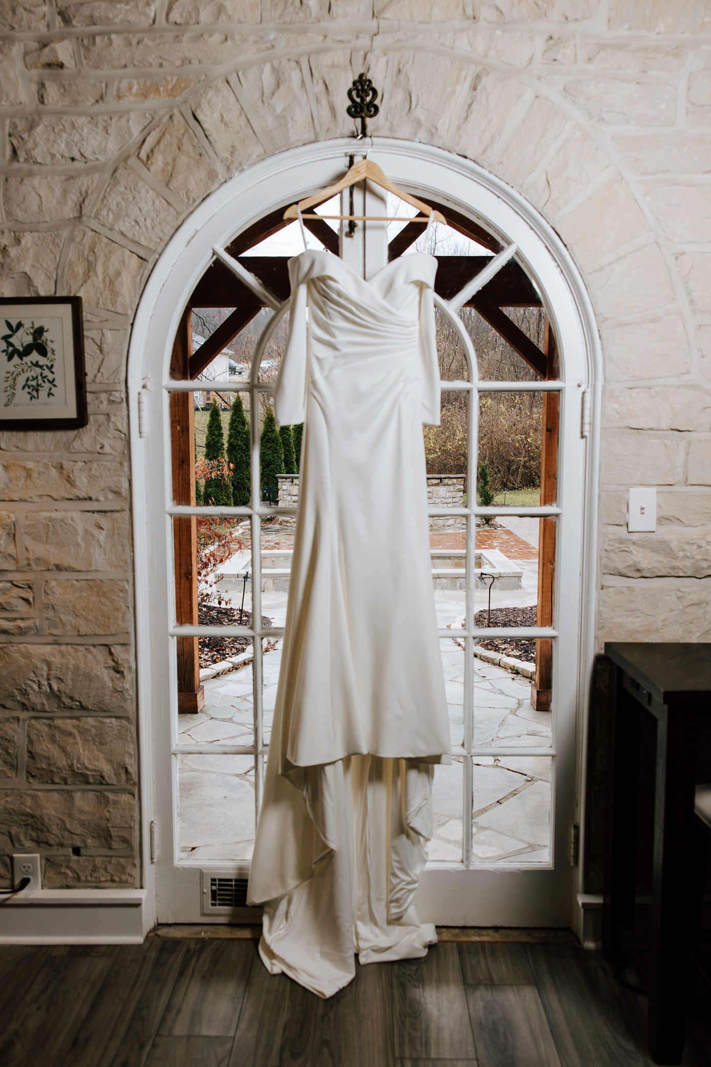 A minimalist white wedding dress with sleeves and ruched fabric hangs from a hook overtop of a doorway in the bridal suite at Knotting Hills in Pevely, Missouri. The walls are made of stone, and a fireplace and garden can be seen in the background through the door's glass window panes.