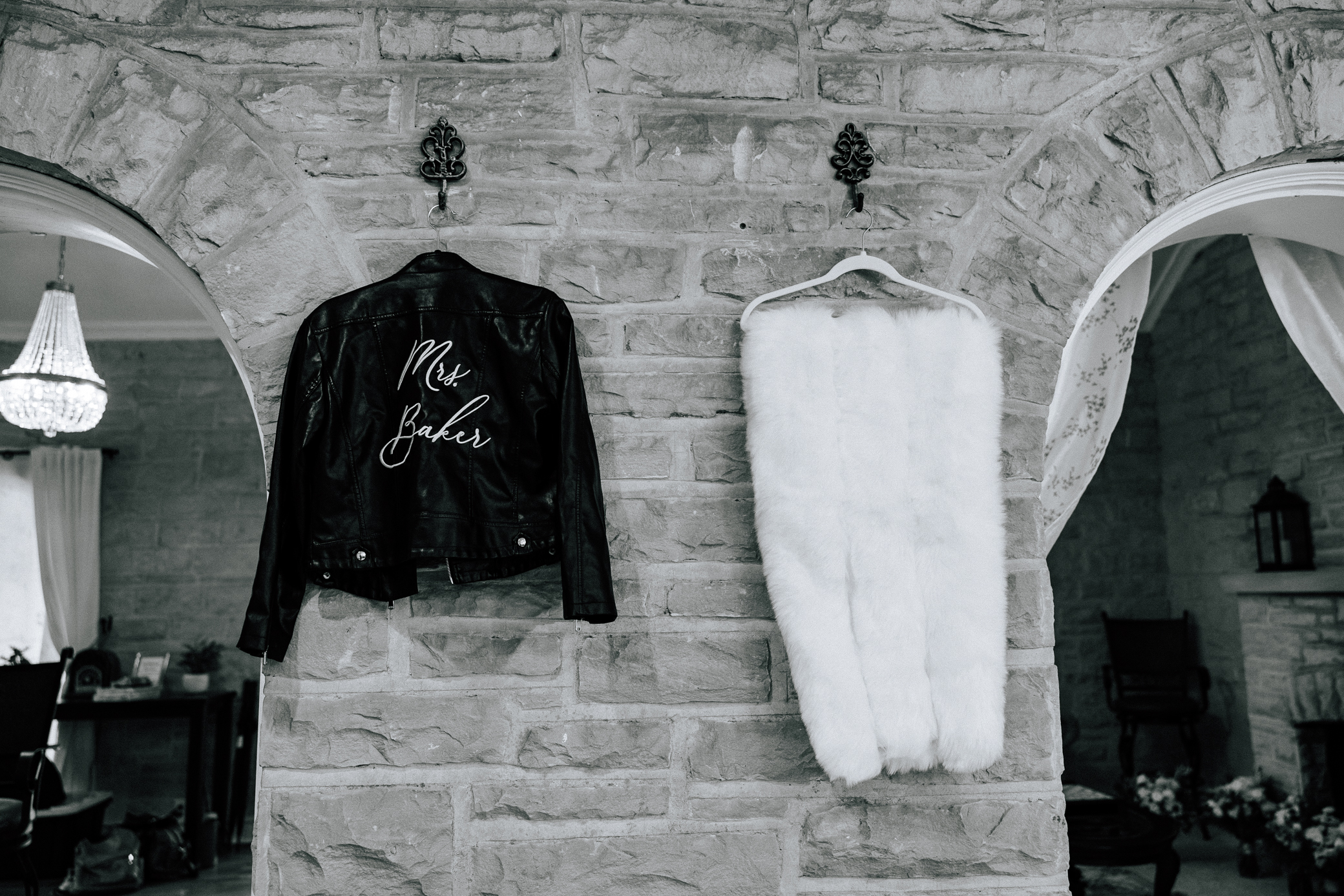 A black and white photo features two important aspects of the bride's attire which can be seen hanging from the stone walls inside of the bridal suite at Knotting Hills in Pevely, Missouri--a white fur shawl to keep warm on a cold day, and a black leather jacket with the words "Mrs. Baker" embroidered on the back in white thread.