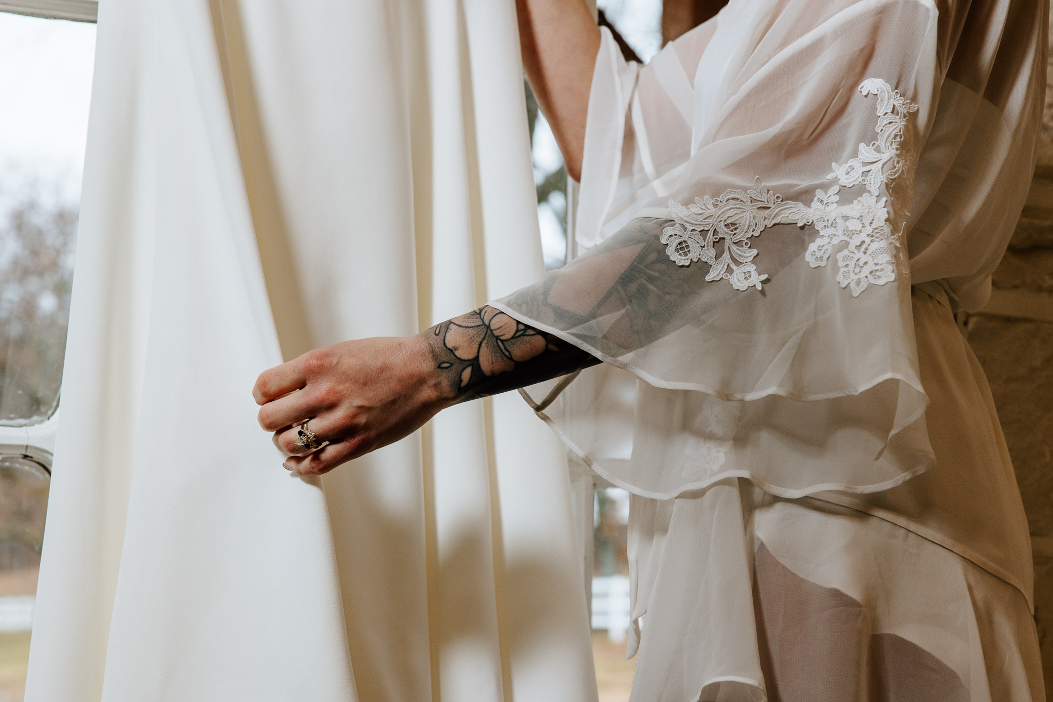 A bride's hand holds the fabric of her dress as it hangs above a window. She's wearing a sheer white lace robe, her black and white diamond engagement ring, and her arm is covered in a floral tattoo sleeve.
