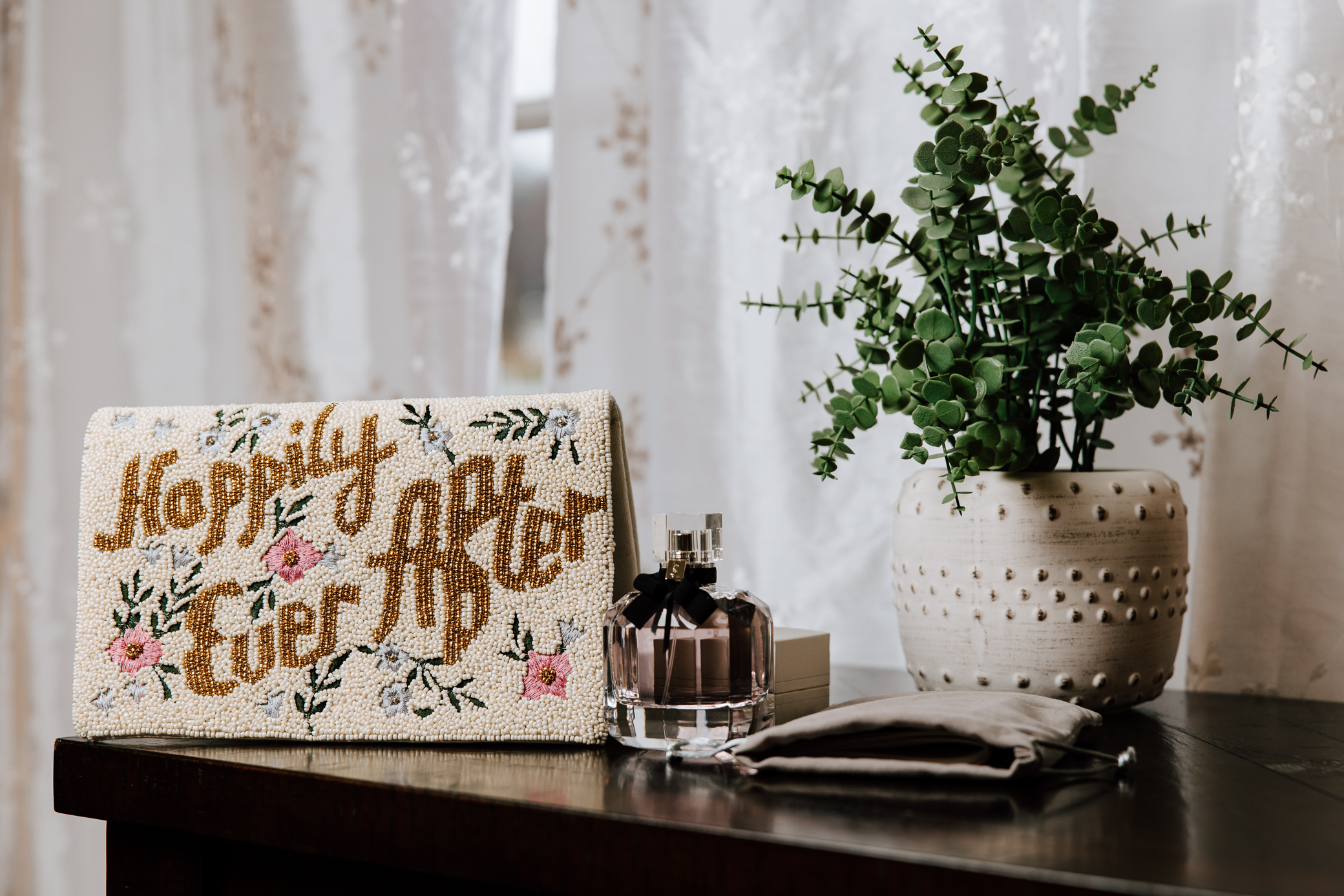 A beaded clutch that reads "happily ever after" is sitting on a table in the bridal suite at Knotting Hills in Pevely, Missouri along with a pink bottle of Yves Saint Laurent Mon Paris perfume and a decorative plant in a white pot.