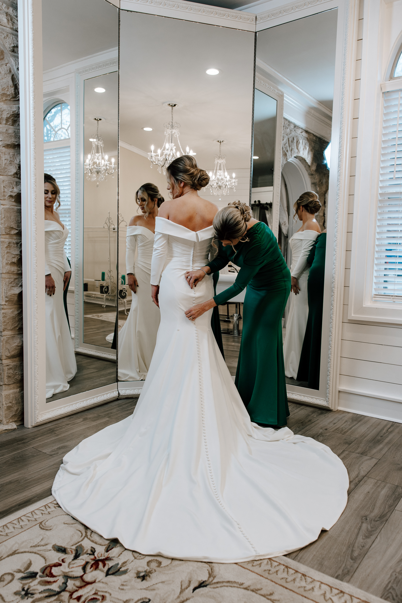 A bride gets buttoned into her white minimalist wedding gown by her mother who is wearing a green velvet gown.