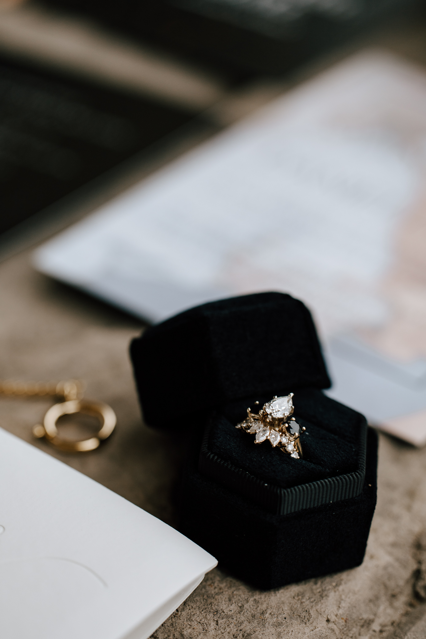 A bride's gold wedding set is shown in a black ring box. The engagement ring features two black diamonds surrounding one large pear cut diamond and the wedding band features several white marquise cut diamonds.