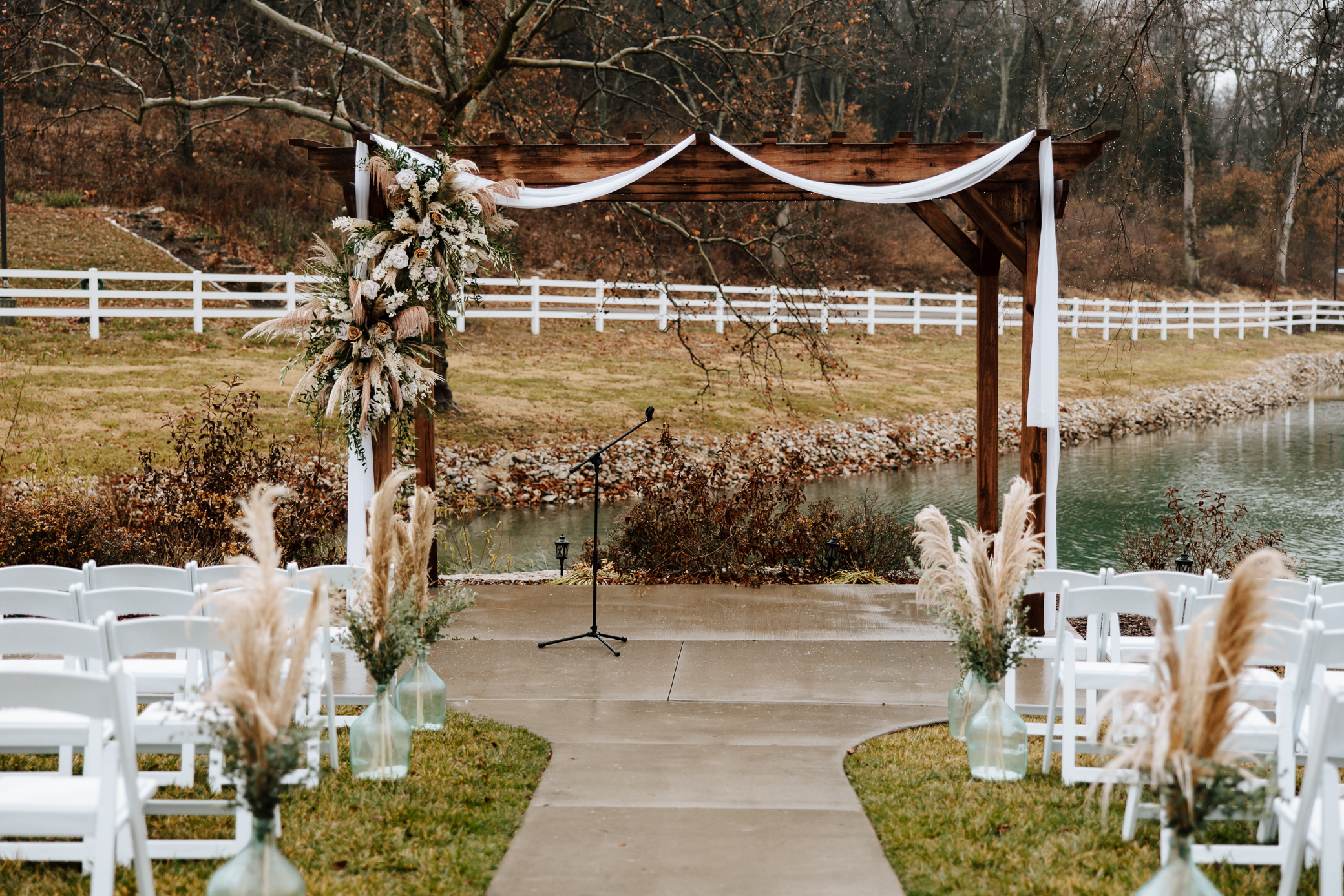 A view of the pergola arbor in the ceremony space at Knotting Hills in Pevely, Missouri. The pergola is adorned with white and brown florals with greenery and pampas grass and features a white cloth draped over the top of the arbor.