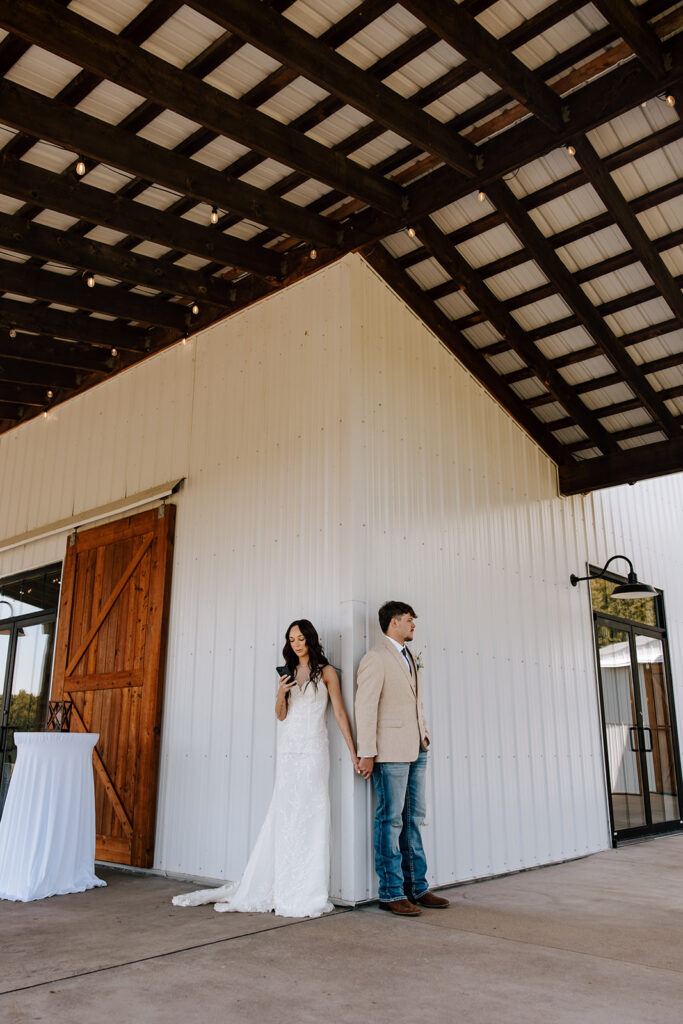 A bride and groom hold hands around a corner of a white barn during their first touch and private vow reading.