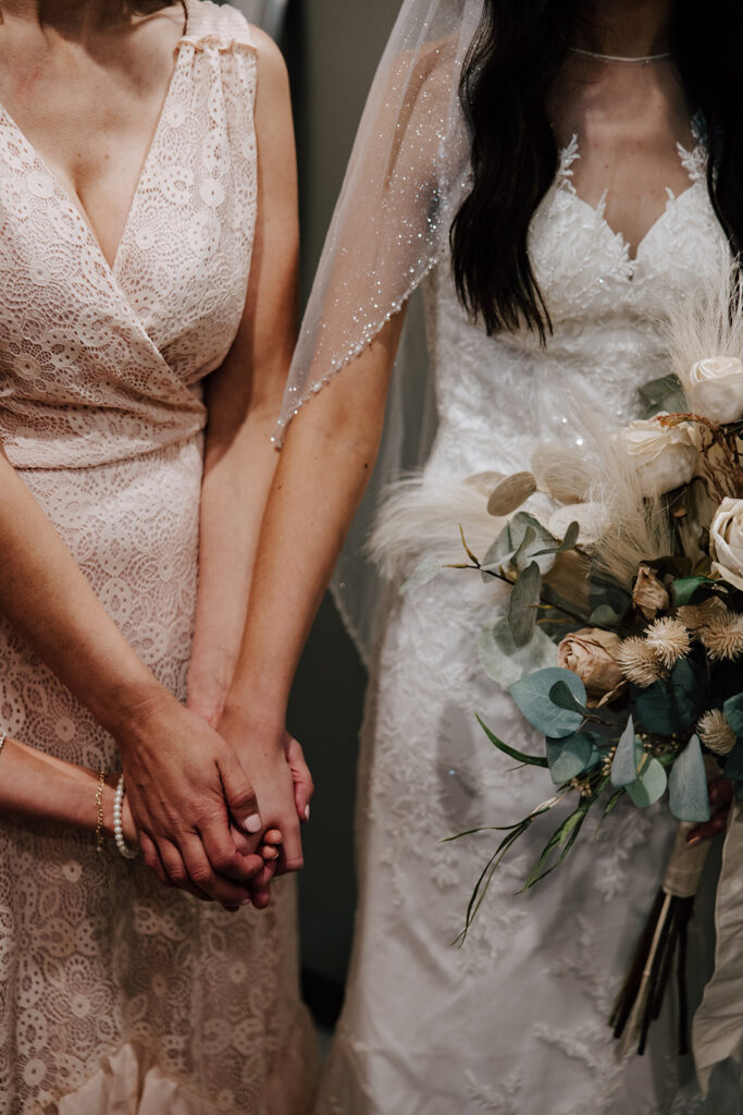 A bride and her mom hold hands during a group prayer before her wedding ceremony. The bride is holding a bouquet of flowers.