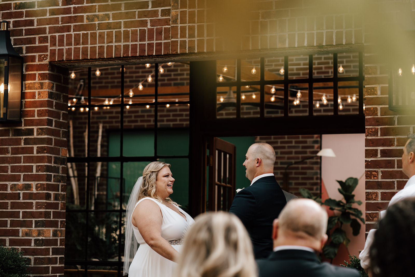 A bride and groom hold hands during their wedding ceremony in front of greenhouse with brick walls.