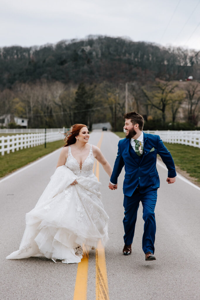 A bride and groom hold hands and smile at each other while running down a road on their wedding day.