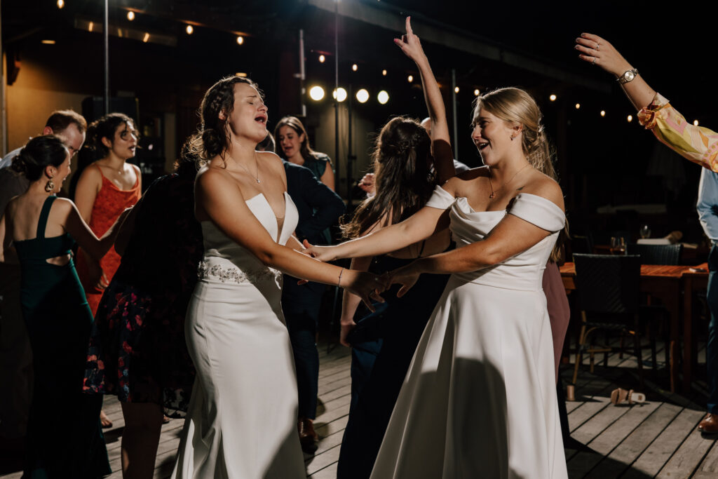 Two brides dance with each other during their wedding reception, surrounded by family and friends.