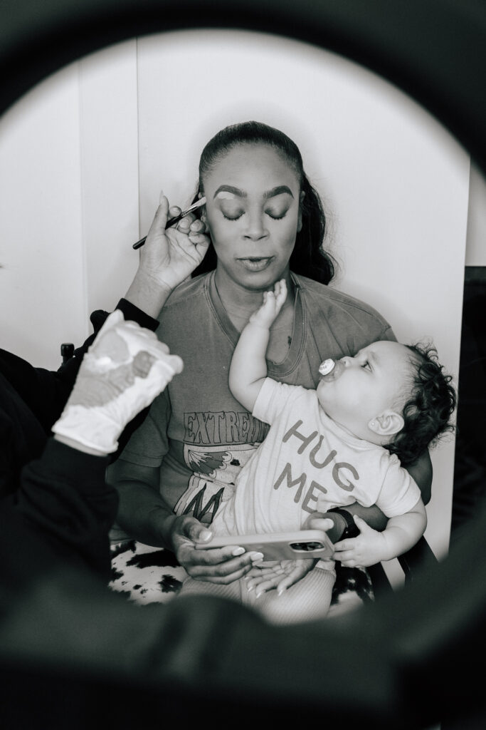 A bride gets her makeup done on her wedding day while her daughter sits on her lap and reaches for her face.