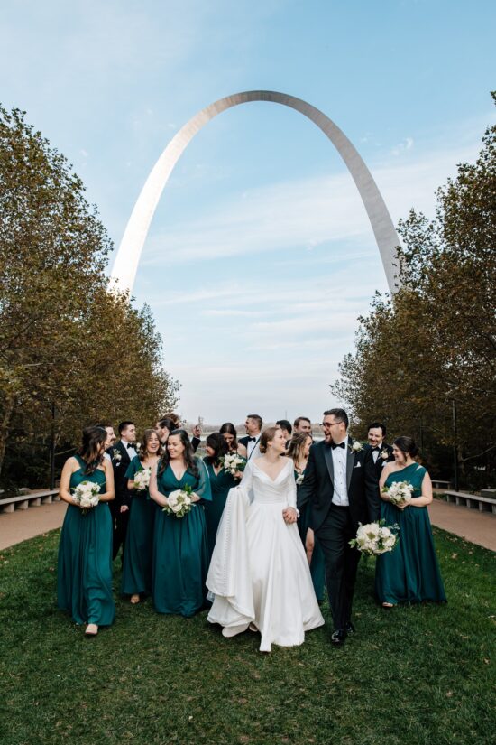 A newlywed couple and their wedding party walk and share some laughs together in front of the Gateway Arch in Gateway Arch National Park in St. Louis, Missouri.