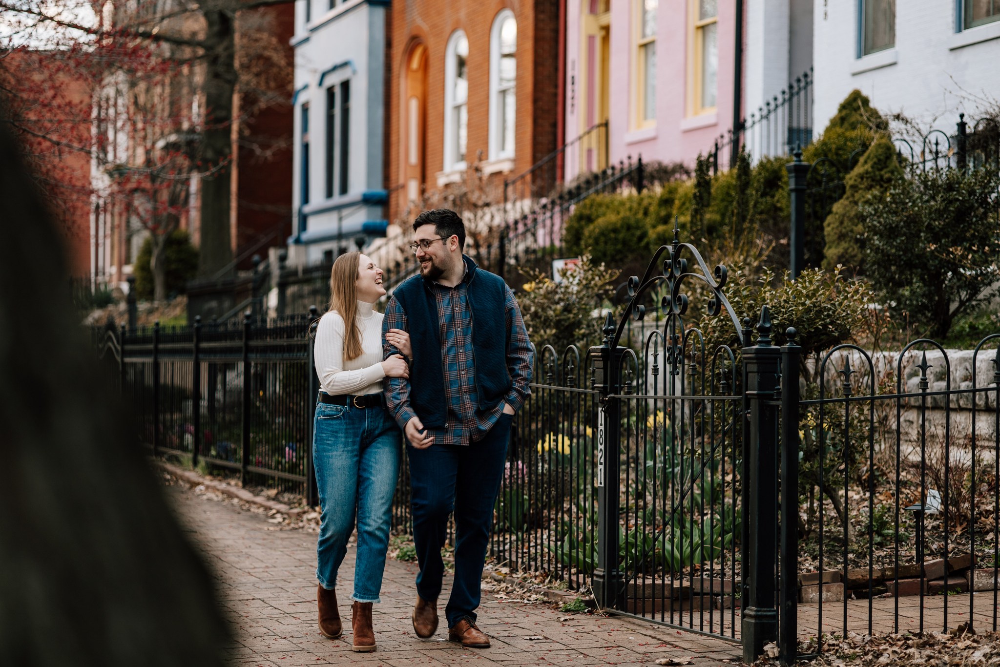 A woman warmly holds onto the arm of a man while walking down a brick sidewalk in front of brightly colored houses and black wrought iron fences. They're dressed warmly and laughing while looking in each others' eyes.