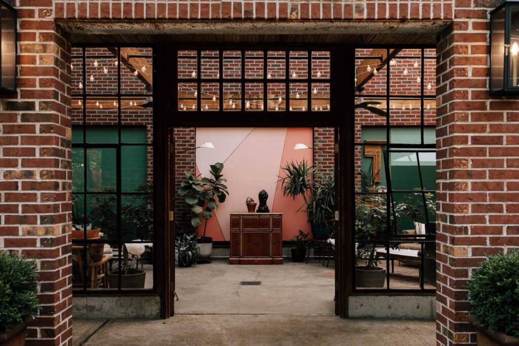 Modern industrial wedding venue featuring exposed brick walls, large glass doors, indoor greenery, string lights, and a statement wall with geometric pastel designs, creating a stylish and unique backdrop.