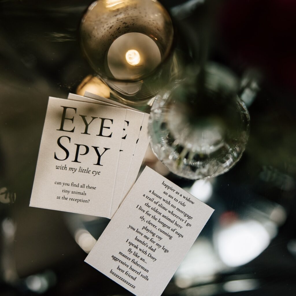 Close-up of 'Eye Spy' wedding reception game cards placed on a reflective glass table, surrounded by candlelight and delicate decor, adding a fun and interactive element to the celebration.