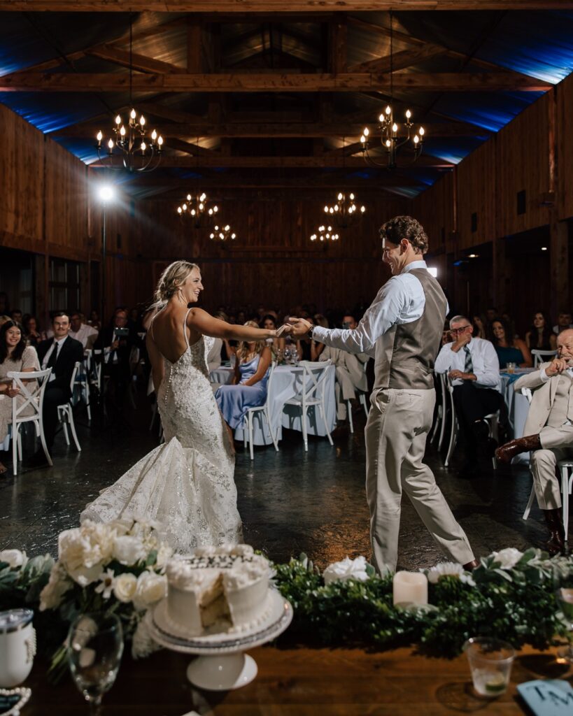 Bride and groom sharing their first dance in a rustic barn wedding venue, surrounded by guests seated at white tables with soft lighting and wooden beams, featuring a beautifully decorated wedding cake and greenery on the foreground table.