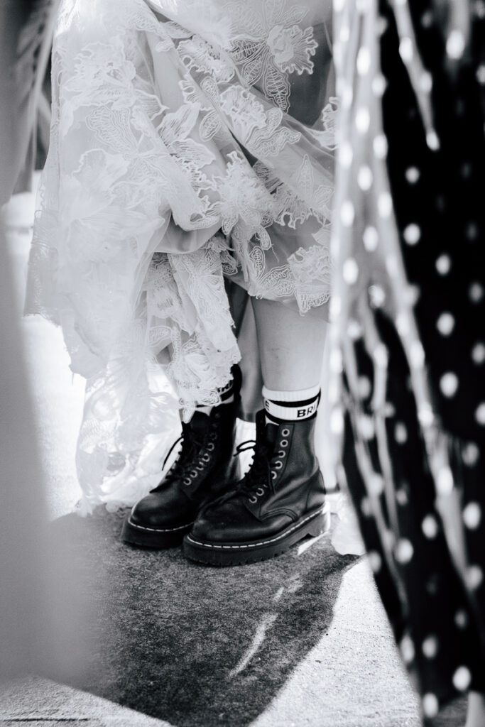 A bride is holding her dress in a bunch to show off her black Doc Martens with socks that say "bride" on the top band. The photo only shows her from the knees down.