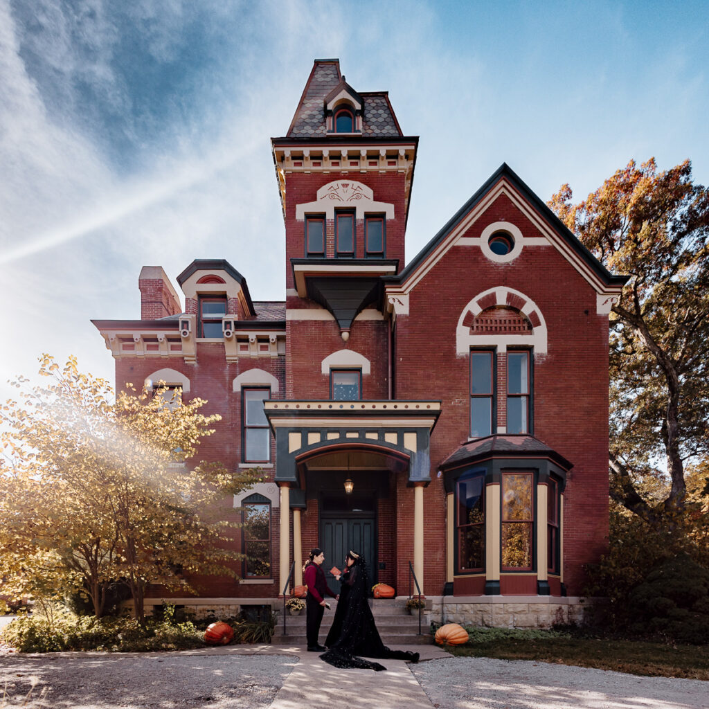 A man and woman wearing black and red hold hands while exchanging wedding vows in front of a rehabilitated Victorian-era home. A tree with yellow leaves and several large pumpkins are seen on and around the house's front porch.