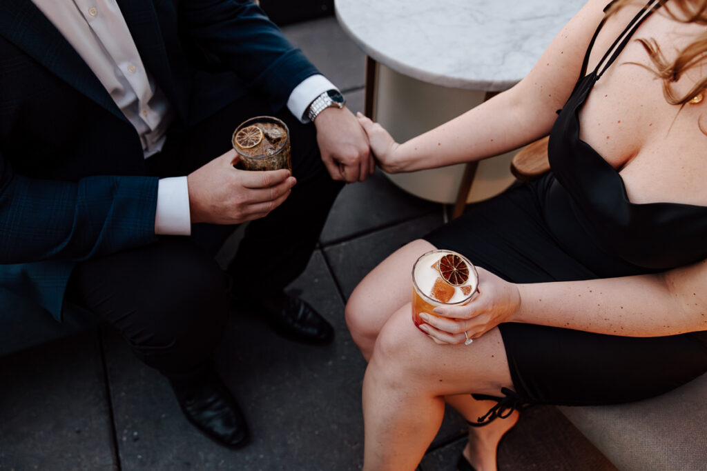 A newly engaged man and woman hold a drink in one hand and hold hands with the other.