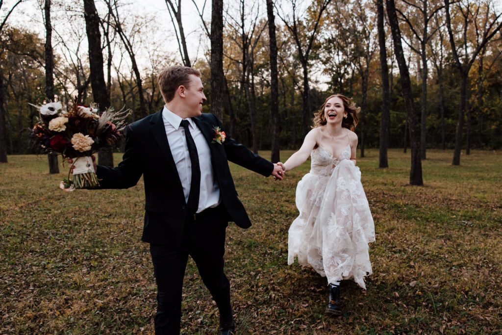 A bride and groom playfully run through a beautiful forested area after getting married.