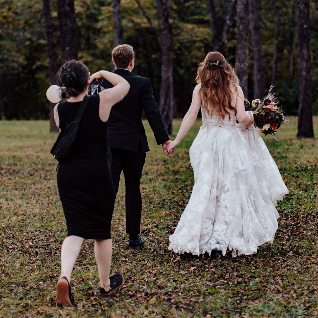 A photographer is pictured taking photos of a newly married couple walking away into a grove of trees.